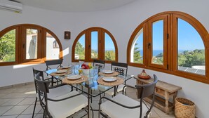 A dining area with a glass table, chairs, and a view of the sea through large arched windows. - Milugar - Javea, Costa Blanca (Javea)
