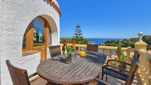 A round wooden table with wicker chairs, a vase with red flowers, and a glass of orange juice on a balcony overlooking the sea. - Milugar - Javea, Costa Blanca (Javea)