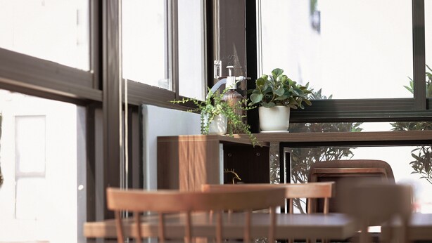Una silla y una mesa de madera en una habitación con ventanas amplias, una planta en maceta y un dispensador de gel hidroalcohólico en el alféizar.