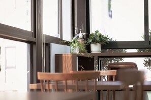 A wooden chair and table in a room with large windows, a potted plant, and a bottle of hand sanitizer on the windowsill. - The Cosy Inn Hanoi Hotel (Hanoi)