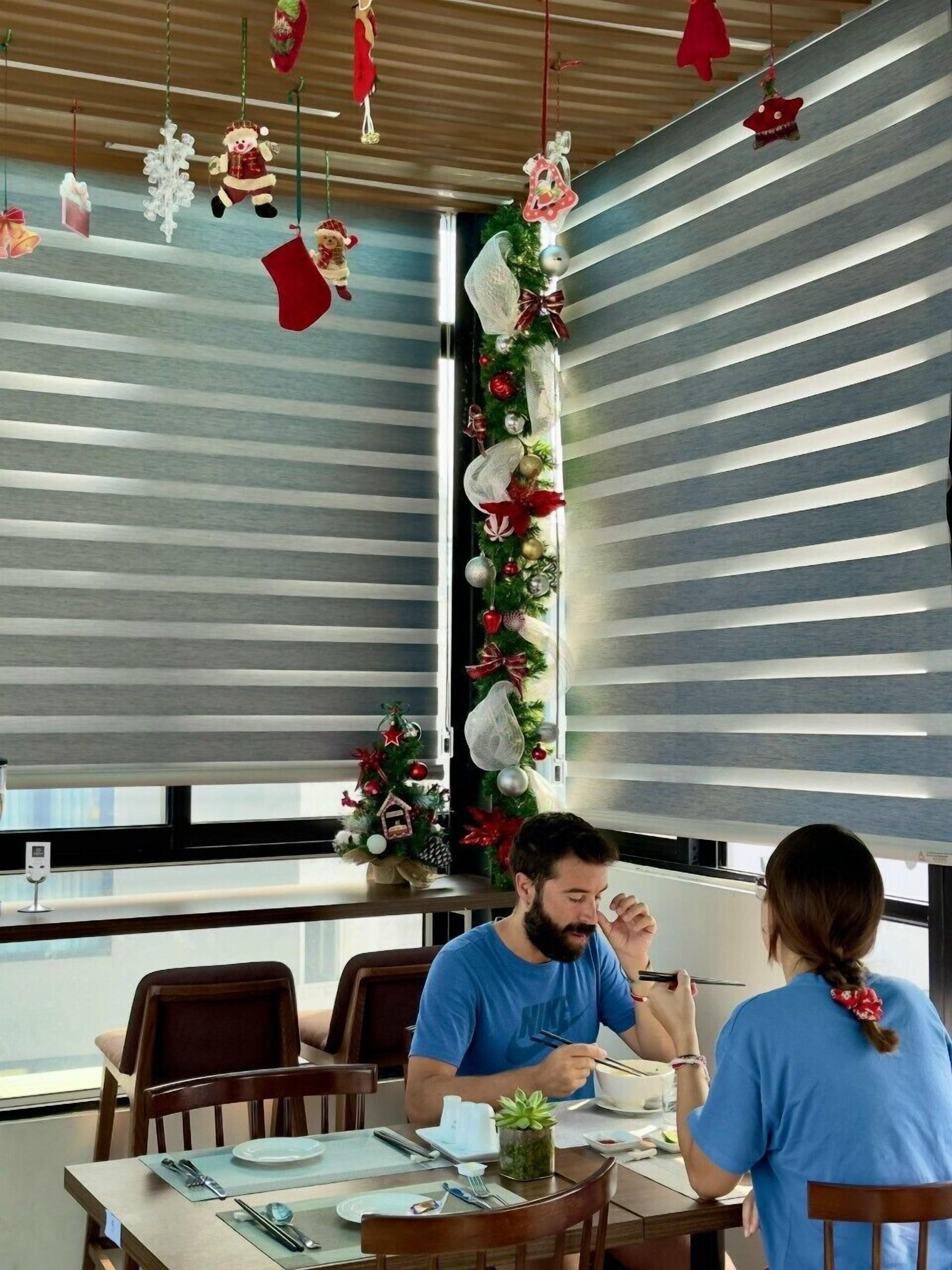 A dining area with a Christmas tree and decorations, a window with blinds, and a table set for two people.