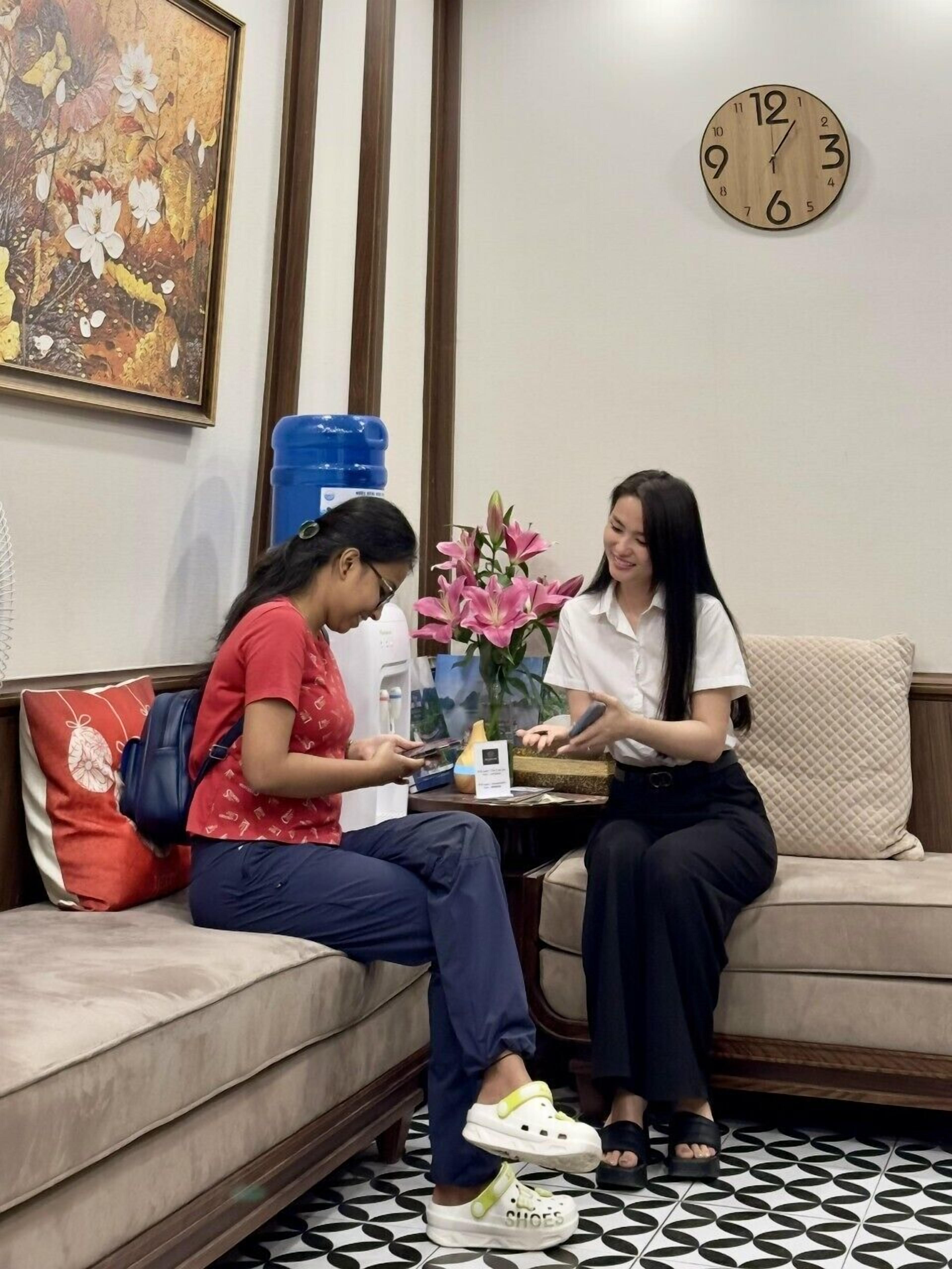 Two people sitting on a sofa in a room with a clock on the wall, a water dispenser, and a vase with flowers.