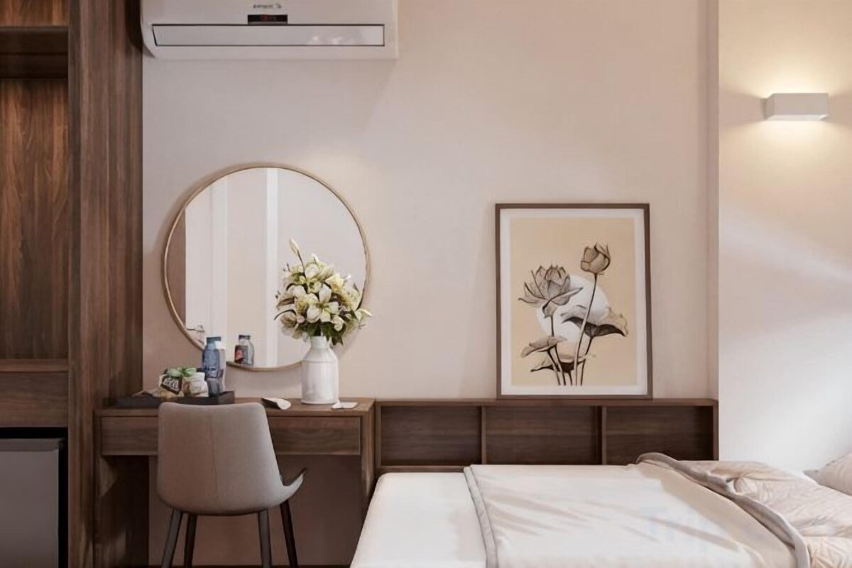 A hotel room with a bed, a desk with a chair, a round mirror, a framed picture, and a vase with flowers.