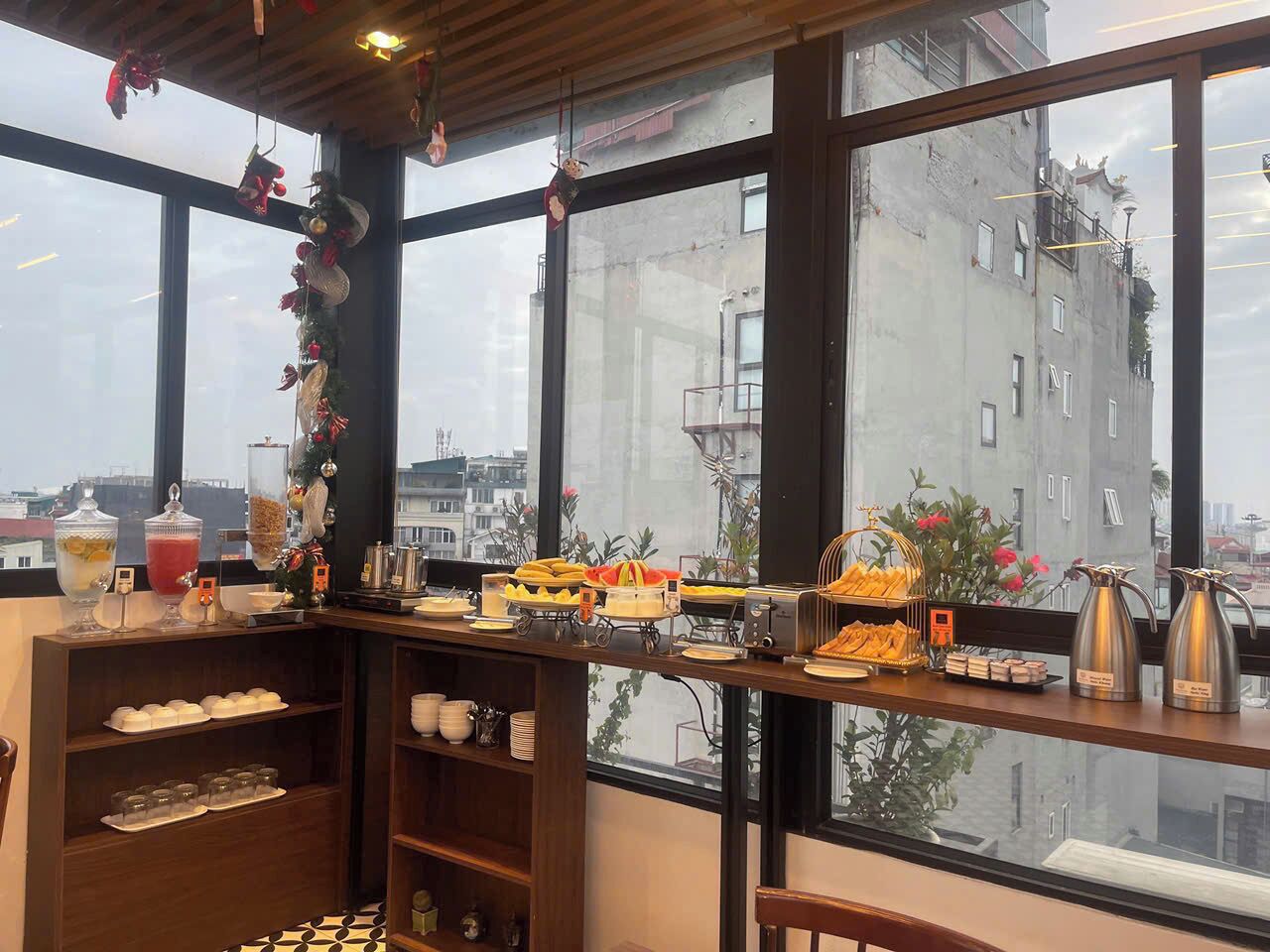 A breakfast buffet with various food items and beverages, a coffee station, and a view of buildings through large windows.