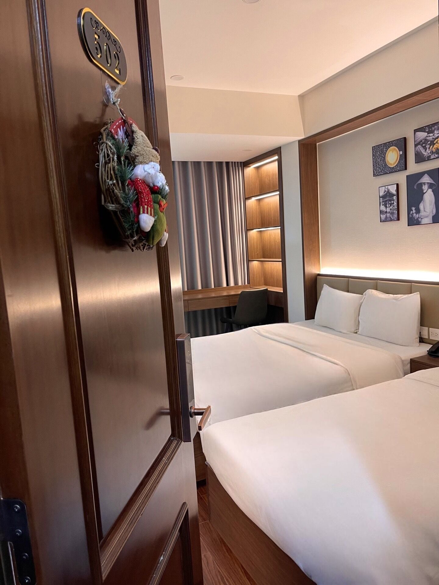 A hotel room with two beds, a wooden door with a number sign, a wall-mounted shelf with framed pictures, and a small desk with a chair.