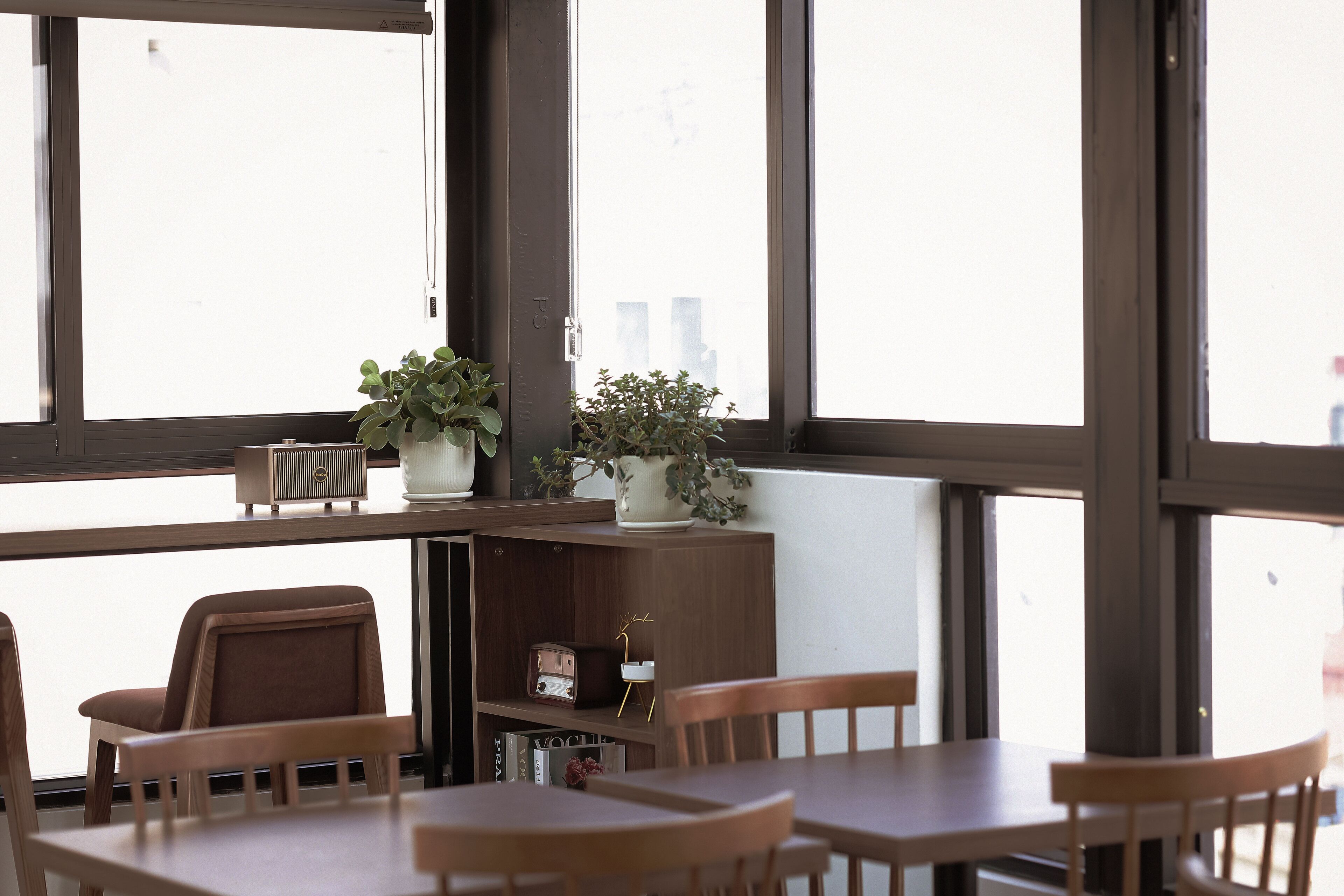 A dining area with wooden chairs and tables, large windows, and a bookshelf with books and decorative items.