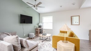 Ein modernes Wohnzimmer mit einer Couch, einem gelben Sessel, einem Couchtisch, einem an der Wand montierten Fernseher und einem Deckenventilator.