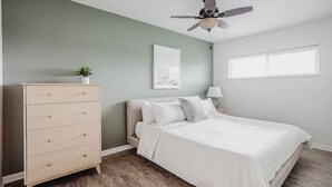 Ein Schlafzimmer mit Bett, Schminktisch, Deckenventilator, Fenster und einem Wandbild.