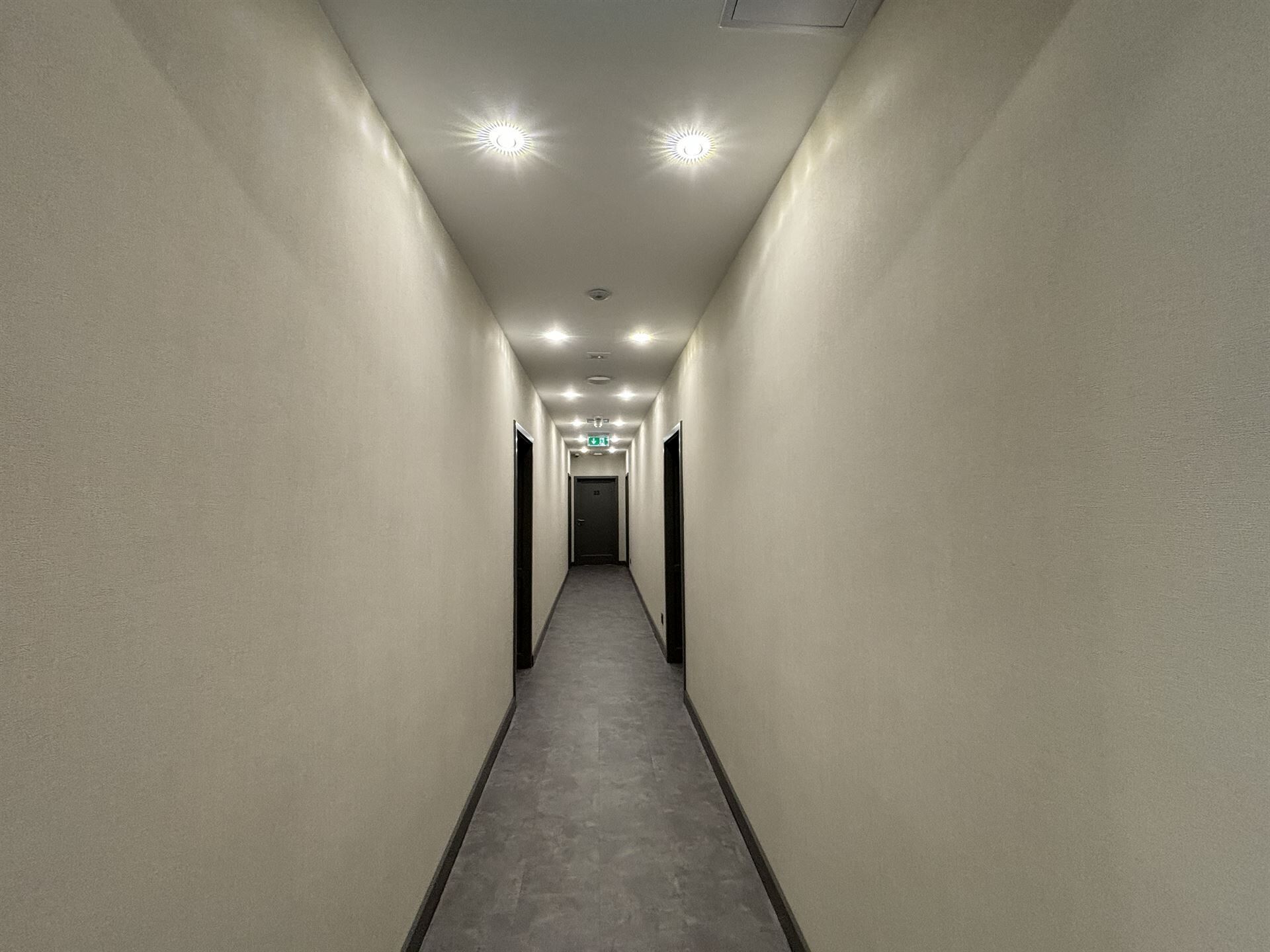 Un couloir étroit et bien éclairé, doté d’un tapis gris et de murs blancs, qui mène à une porte au fond.