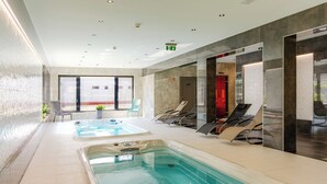 Un área moderna de piscina interior con dos jacuzzis, sillones de descanso y un patio con paredes de vidrio.