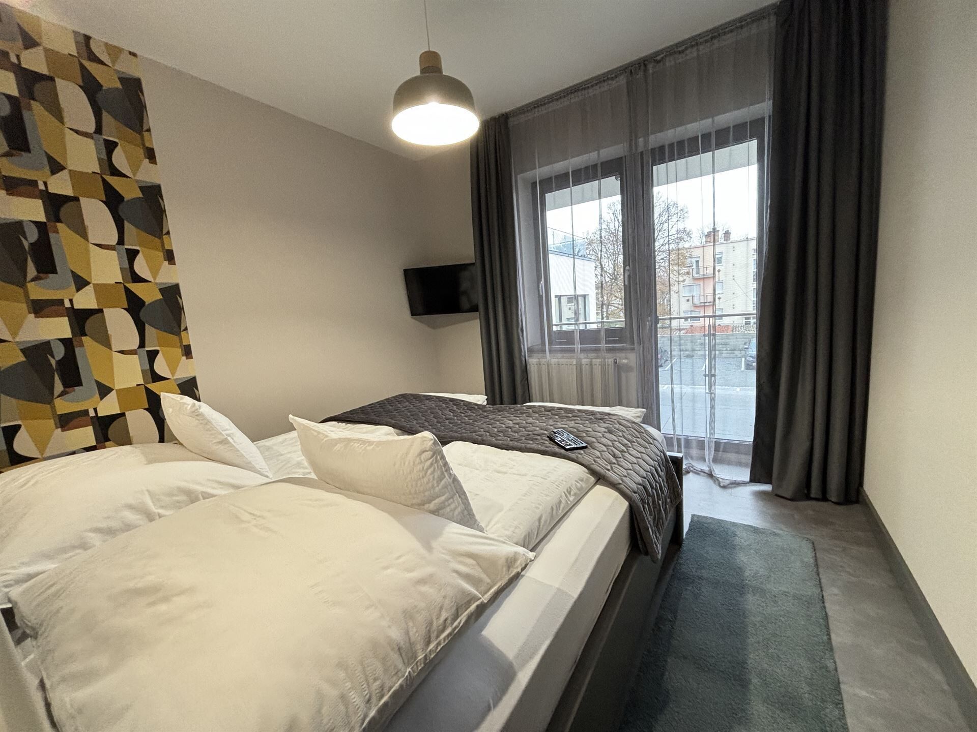 A hotel room with a bed, a TV, a window with curtains, and a geometric wall pattern.