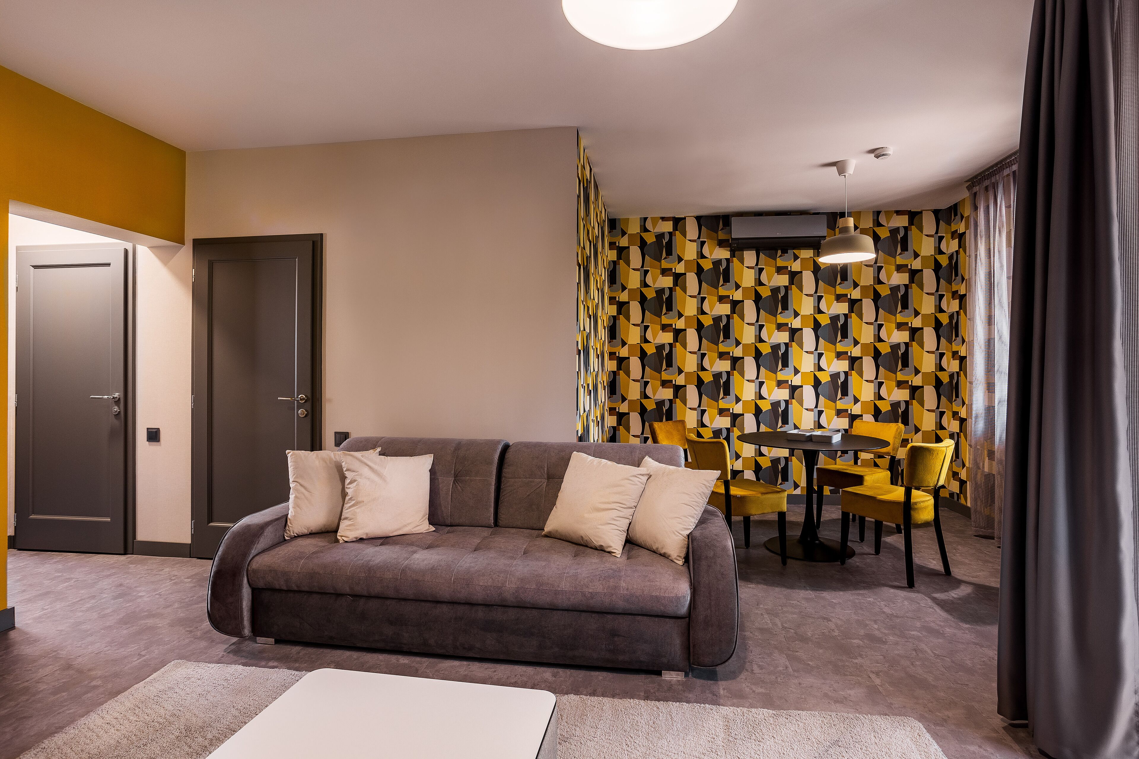 A modern living room with a sofa, a dining table, and chairs. There are two doors and a patterned wall in the background.