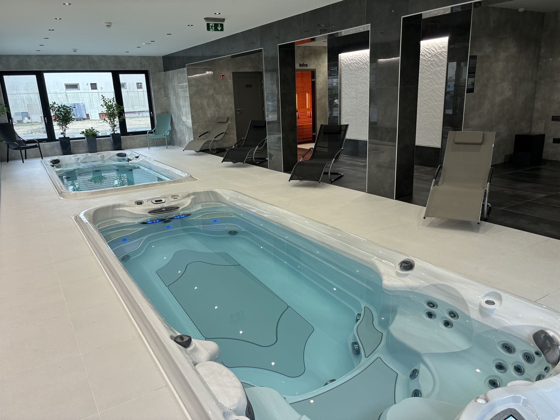 Un área interior de piscina moderna con dos jacuzzis grandes, sillas de descanso y vistas al exterior.