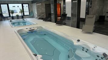 Un área interior de piscina moderna con dos jacuzzis grandes, sillas de descanso y vistas al exterior.