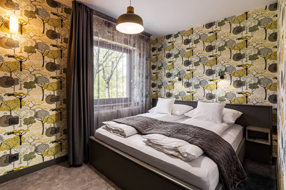 A bedroom with a bed, a window with curtains, and a wall with a tree pattern.