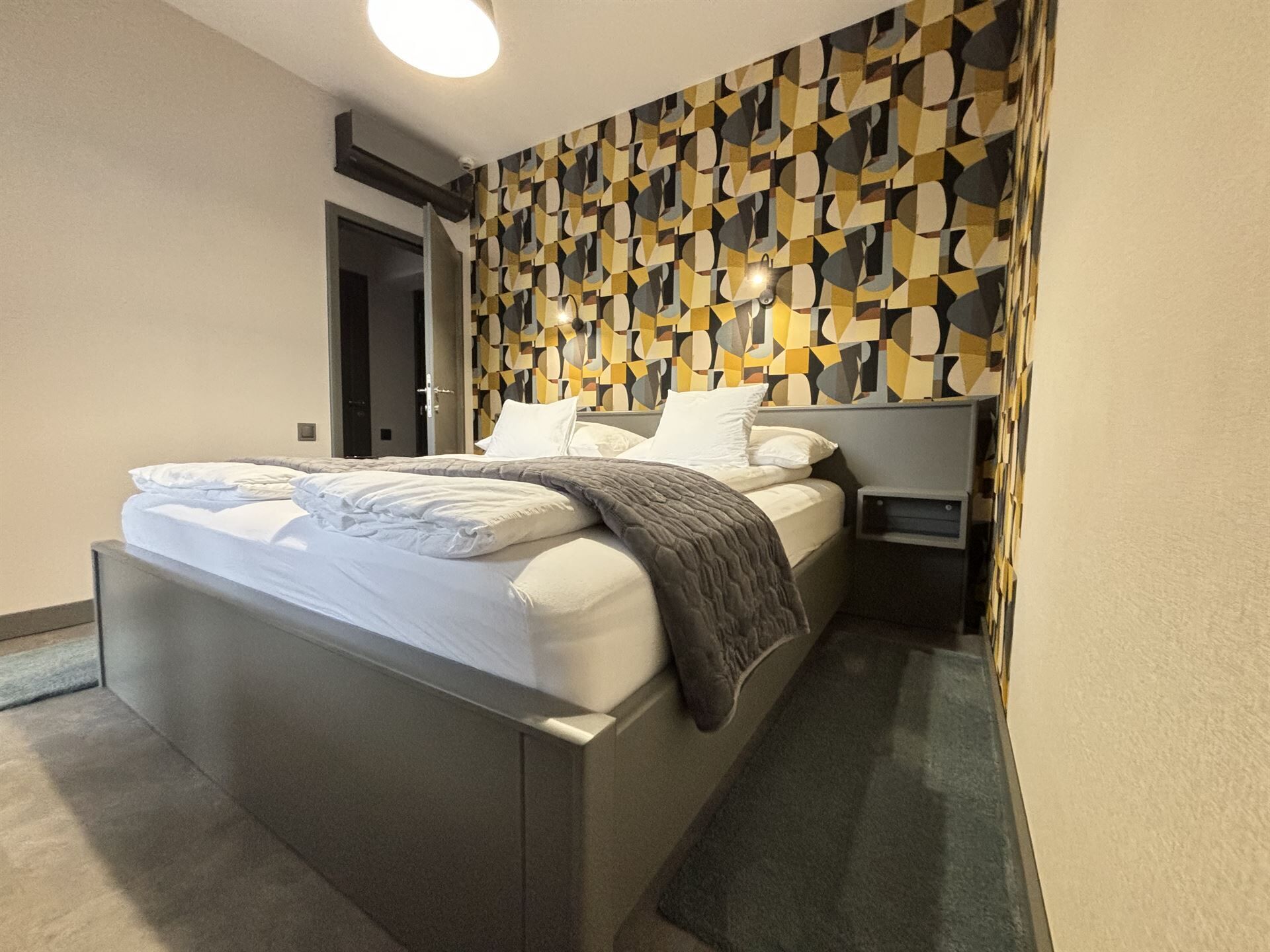 A modern hotel room with a large bed, a bedside table, and a geometric patterned wall.