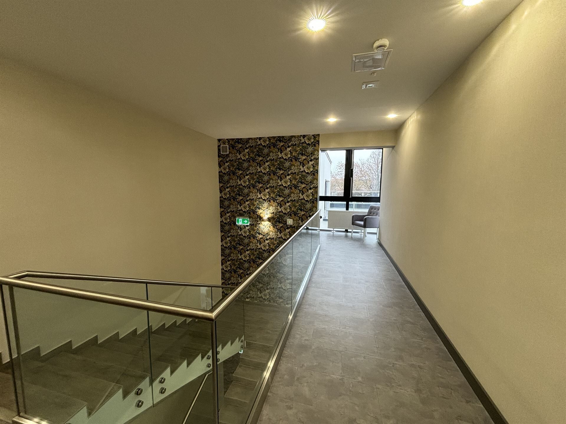 Een modern interieur met een wenteltrap met glazen balustrade, een patroonwand en een uitzicht op een stoel en raam.