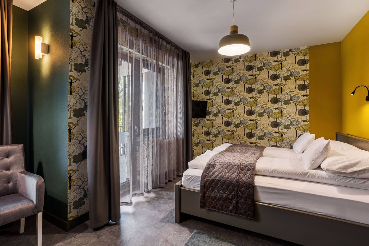 A bedroom with a bed, a chair, a wall-mounted TV, and a window with curtains.