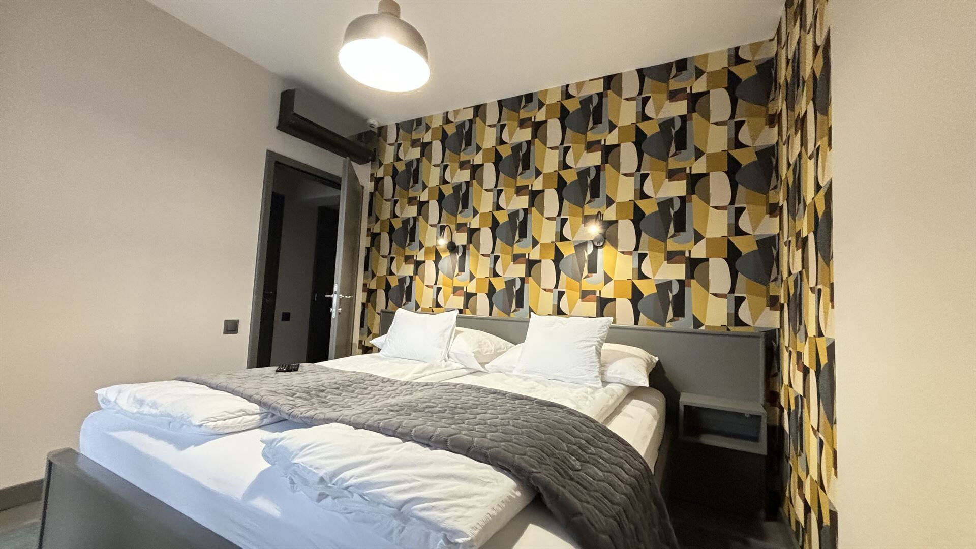 A modern bedroom with a geometric patterned wall, a bed with white linens, and a bedside table.