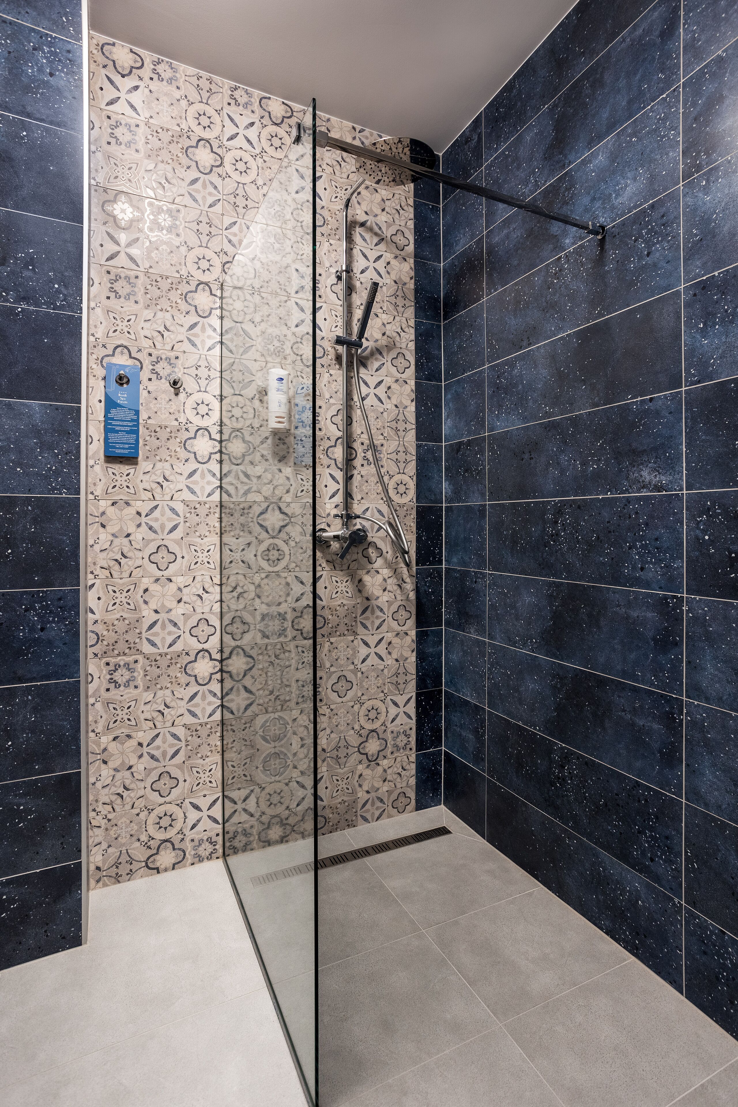 Zona de ducha con mampara de vidrio, ducha de mano y pared con diseño de baldosas geométricas.