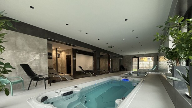 Un área interior de piscina moderna con una gran piscina rectangular, sillas de descanso y un espacio de estar con sillas y una mesa.