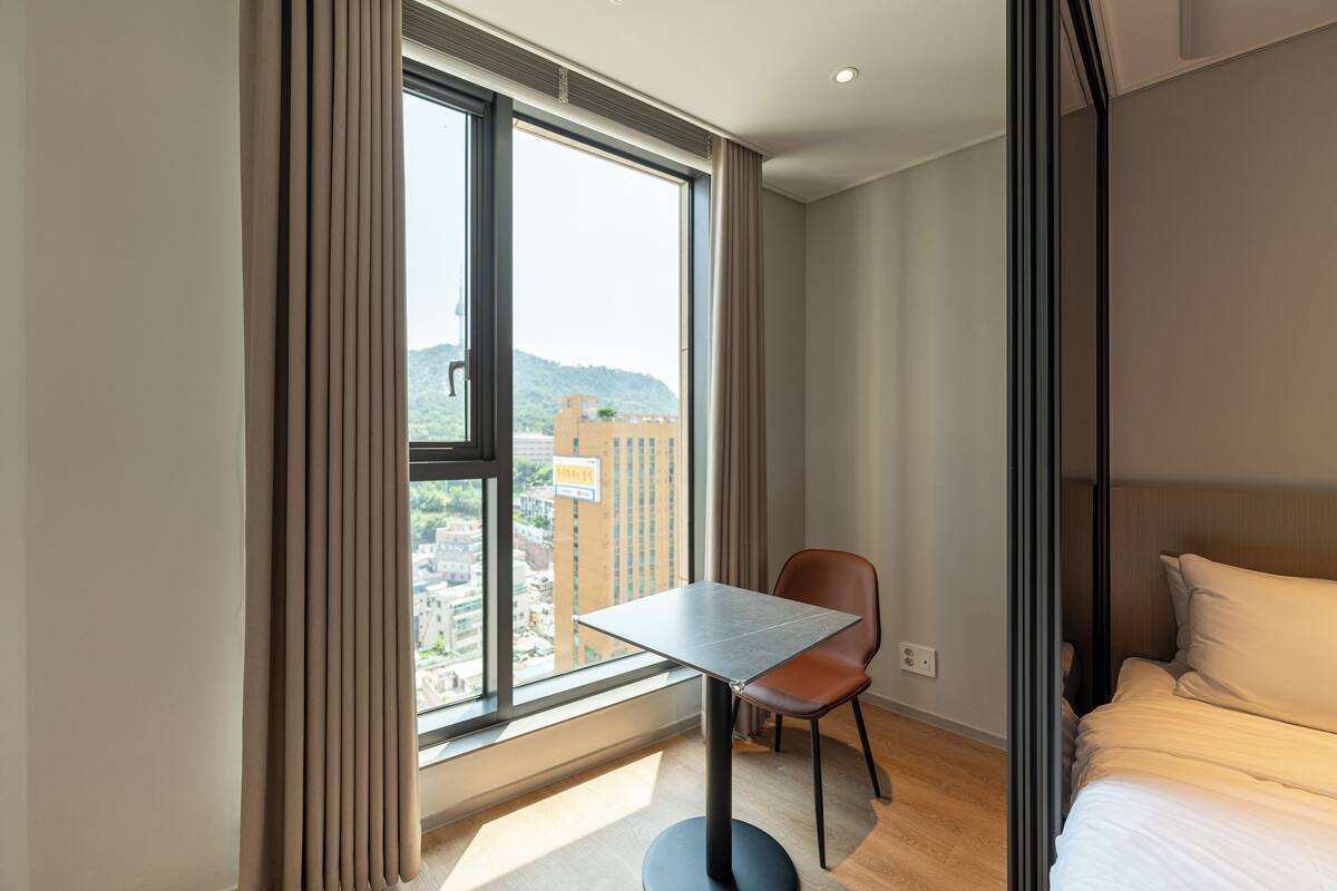 A hotel room with a large window, a single bed, a small table, and a chair.