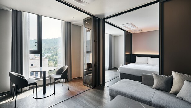 Una habitación de hotel moderna con una cama grande, un sofá, una mesa de comedor con sillas y un ventanal con vistas a la ciudad.