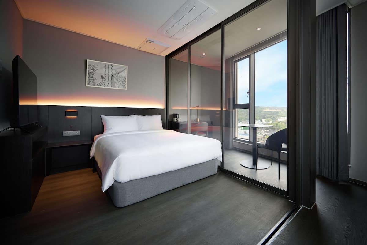 A modern hotel room with a large bed, a bedside table, a nightstand, a desk, and a view of the city through floor-to-ceiling windows.
