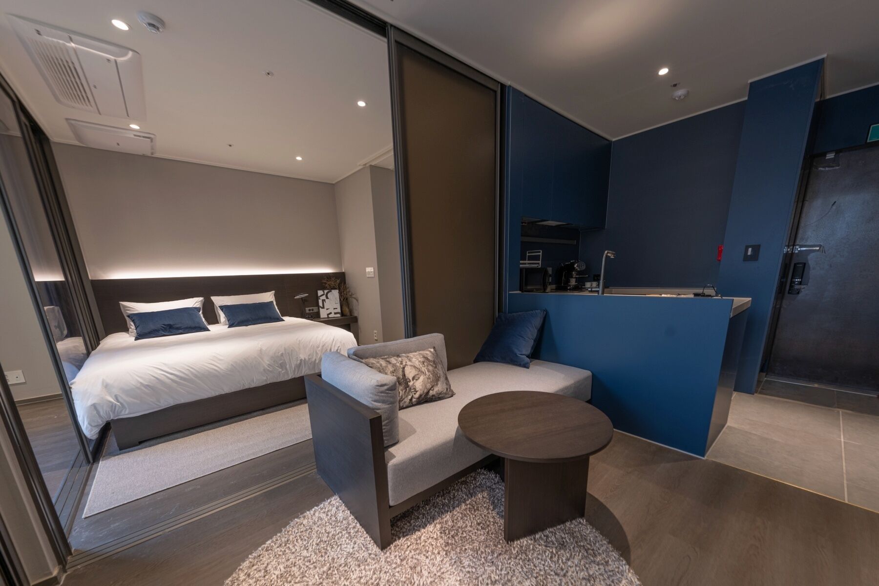 Una habitación de hotel moderna con una cama grande, un sofá, una mesita y una kitchenette.