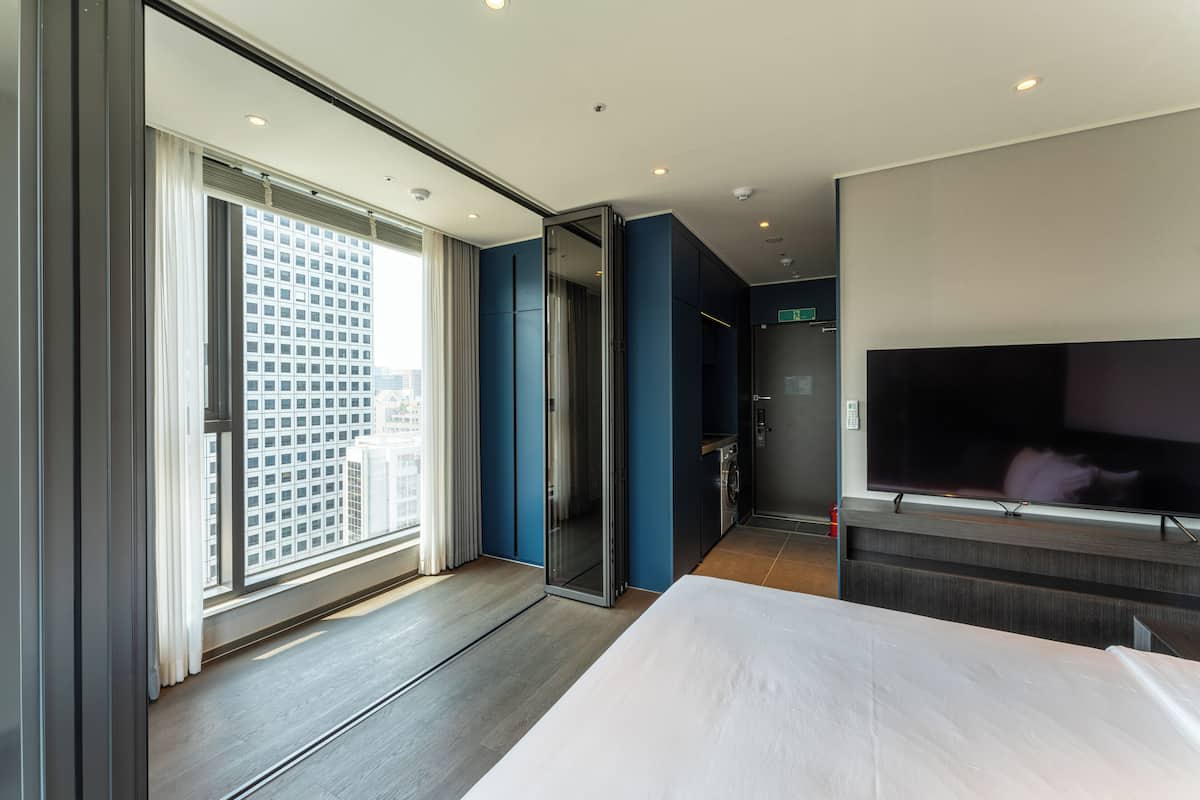 A modern hotel room with a large bed, a flat-screen TV, a sliding door leading to a balcony, and a city view.