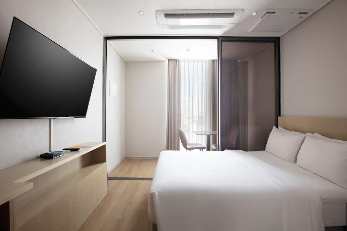 A modern hotel room with a flat-screen TV, a desk, a bed, and a sliding door leading to a balcony.