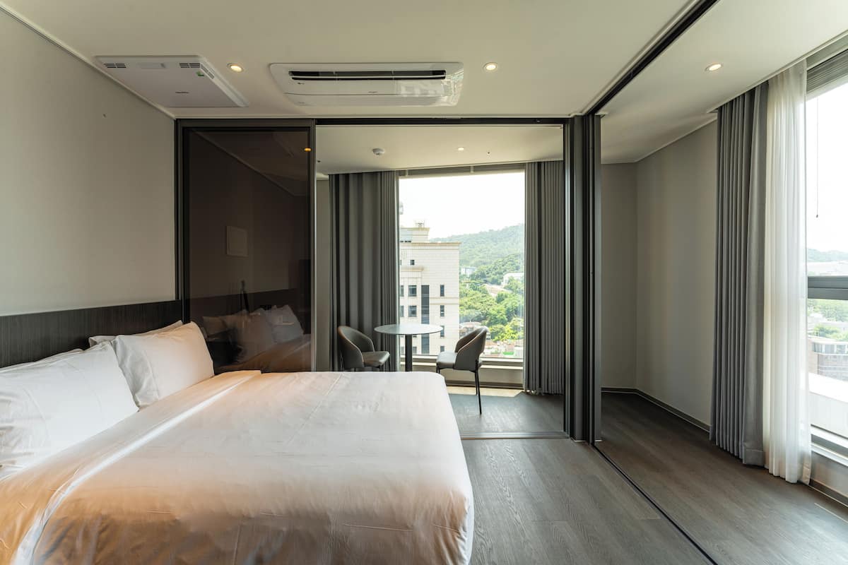 A modern hotel room with a large bed, a small table, and a balcony with a view of buildings and greenery.