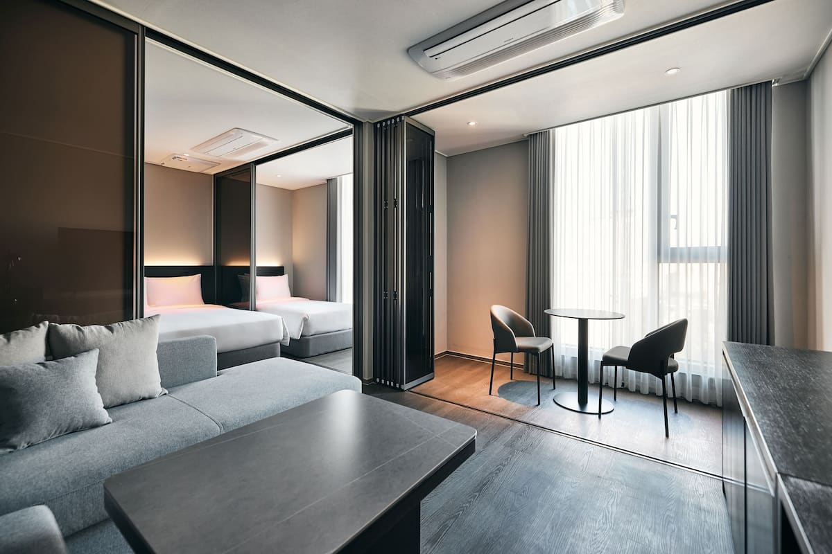 A modern hotel room with a sofa, a dining table, a small kitchen area, a bed, and large windows with curtains.