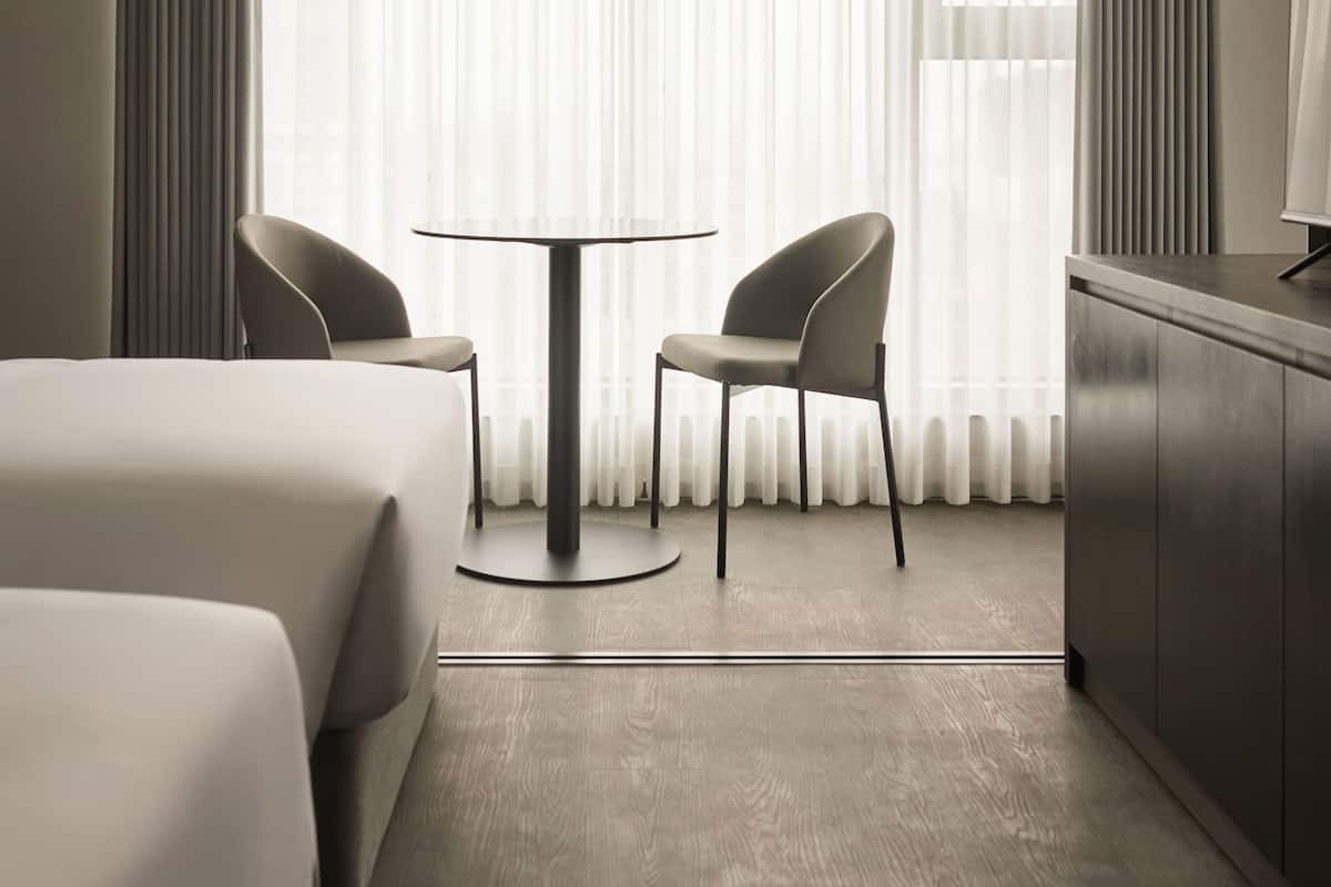 A hotel room with two beds, a round table, two chairs, a TV, and a large window with sheer curtains.