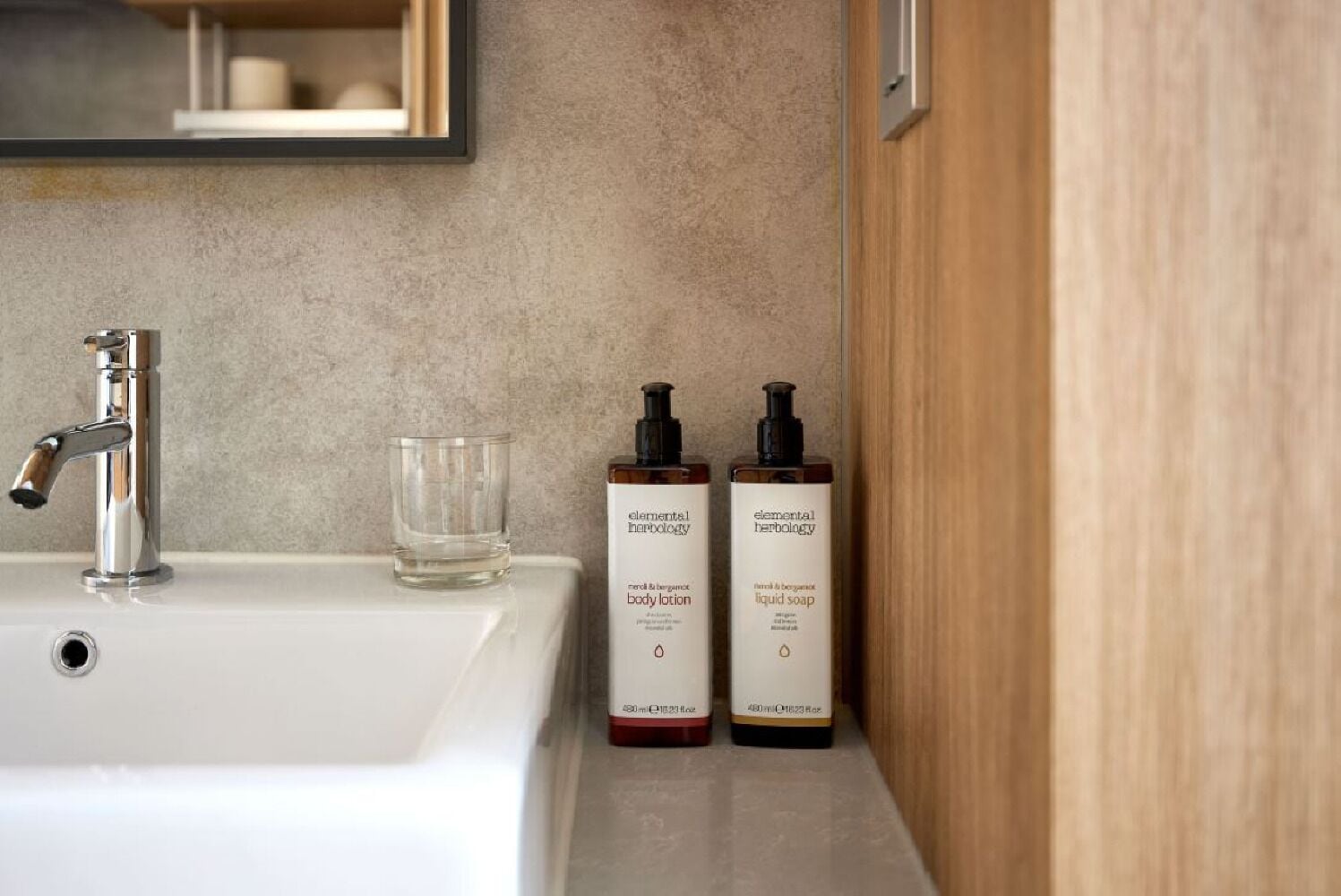 A bathroom sink with a mirror above it, two bottles of Elemental Herbology body lotion and liquid soap, and a glass of water.
