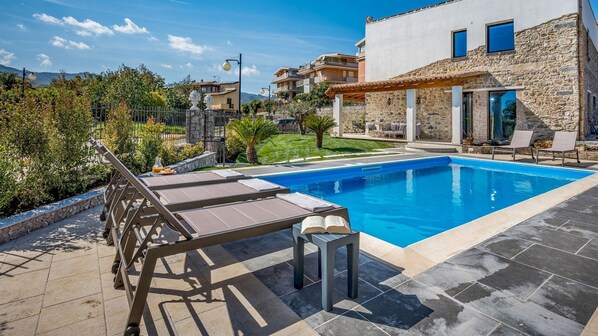 A swimming pool with lounge chairs, a stone wall, and a view of surrounding buildings.