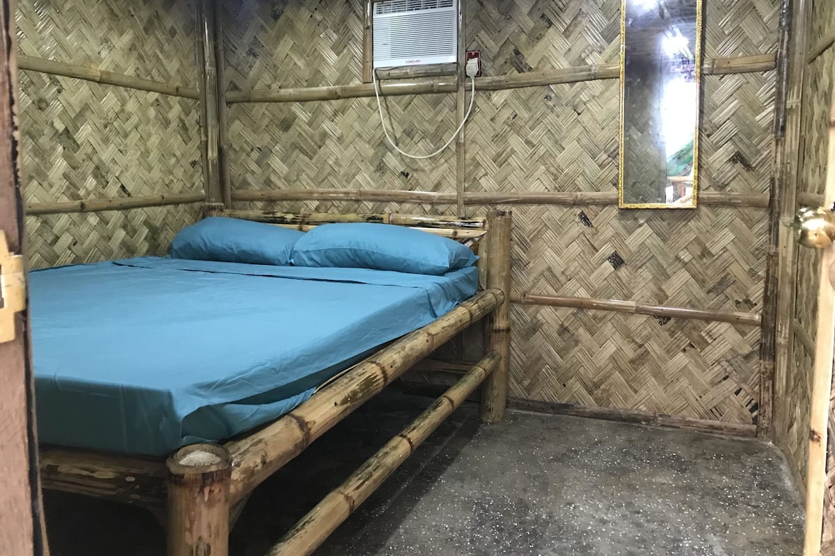 A room with a bamboo wall, a bed with blue bedding, a small mirror, and a ceiling fan.