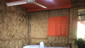 Una habitació amb un llit, un ventilador de sostre, una finestra amb cortina taronja i una taula petita amb una planta.