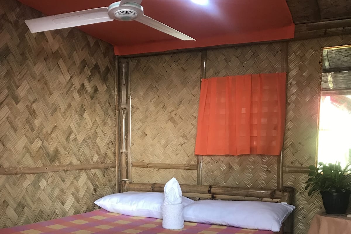 A room with a bed, a ceiling fan, a window with an orange curtain, and a small table with a plant.
