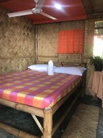 A room with a bed, a ceiling fan, a window with an orange curtain, and a small table with a plant.