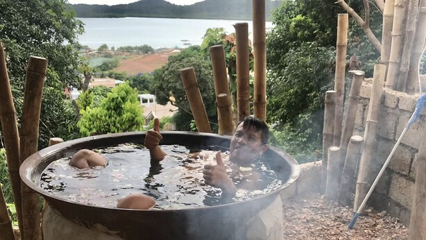 Jacuzzi amb una persona fent el polze en alt, envoltat de canyes de bambú i vista a un llac i les muntanyes.