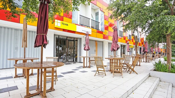 An outdoor café area with wooden tables and chairs, a yellow sign with "TO.HA", and a building with colorful walls in the background. - OYO 92317 Apartment Riverview By Alima View (Cikarang)