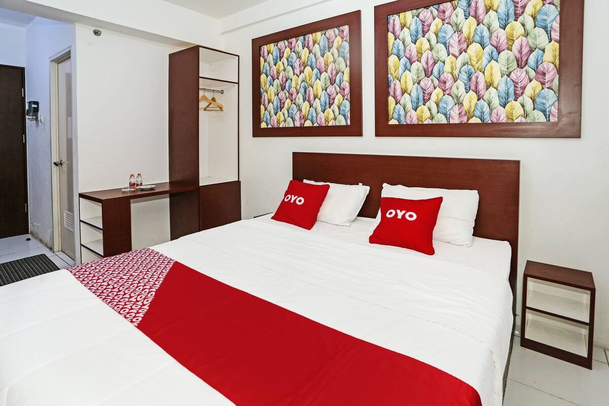 A hotel room with a large bed, two red pillows with the word "OYO" on them, a red and white patterned throw, and two colorful leaf-patterned wall art pieces.
