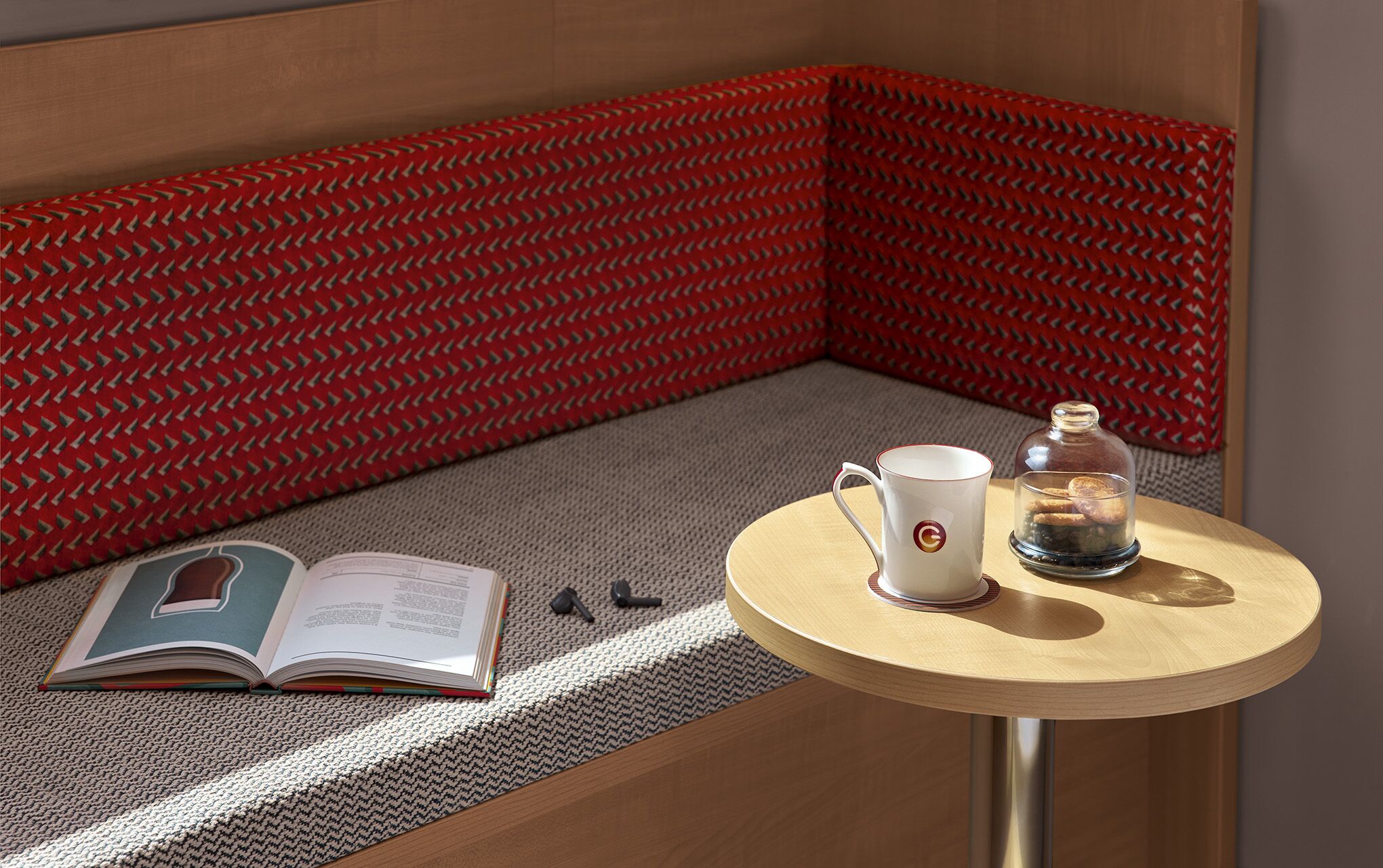 A cozy reading nook with a cushioned bench, a small wooden table with a cup and a glass jar, and an open book.