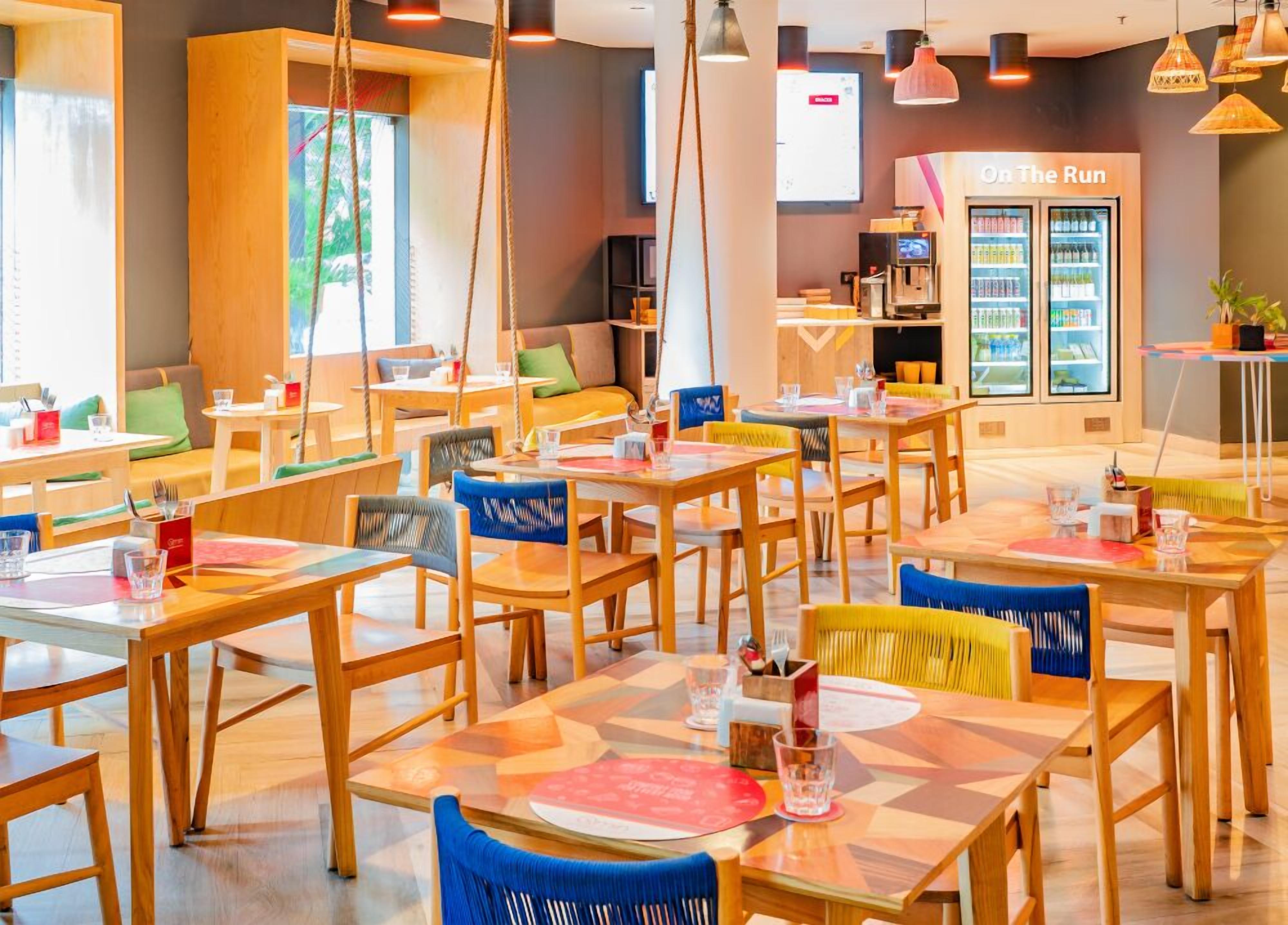 A modern cafe with wooden tables and chairs, a seating area with cushions, and a refrigerator labeled "On The Run" in the background.