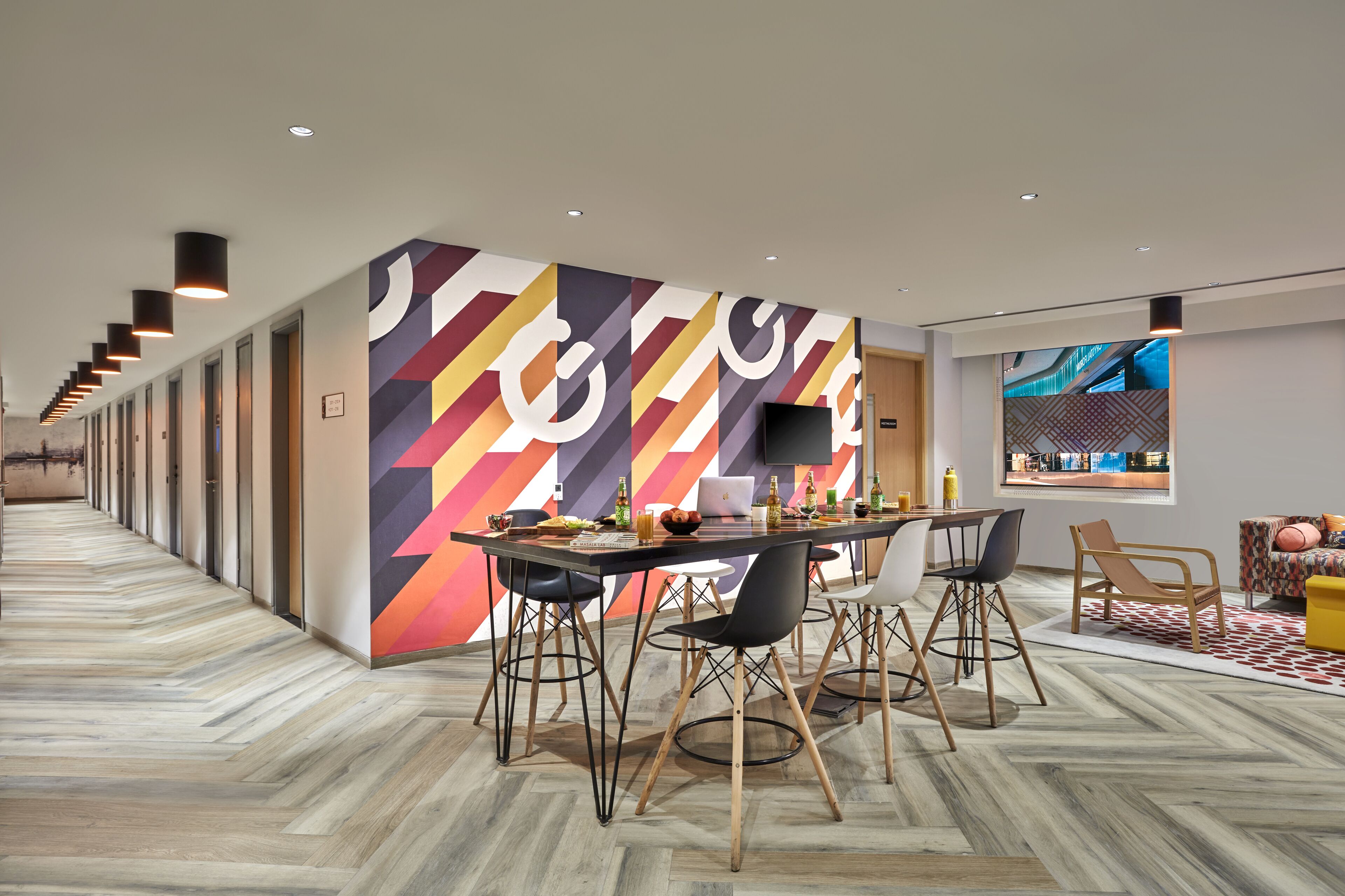 A modern hotel lobby with a long hallway, a dining area with a table and chairs, and a large screen displaying a cityscape.