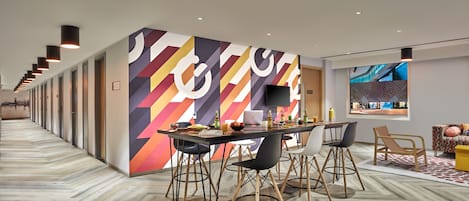 Eine moderne Hotelhalle mit einer langen Korridor, einem Essbereich mit Tisch und Stühlen sowie einem großen Bildschirm, der eine Stadtansicht zeigt.