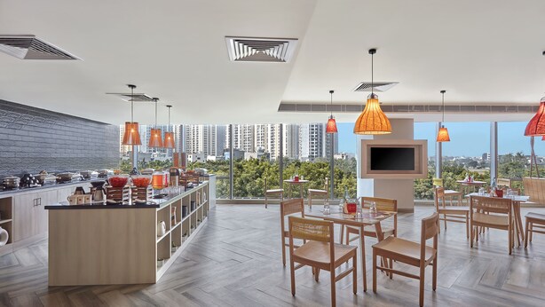 A modern kitchen with a central island, dining area with wooden chairs, and a large TV screen. - Ginger Noida Sector 133 (Noida)