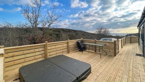 En träveranda med en madrass, ett bord och stolar, med utsikt över ett kuperat landskap.