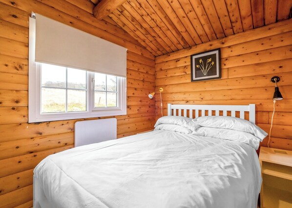 A wooden cabin bedroom with a bed, a window, a framed picture, and a lamp.